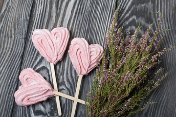 Delicate pink marshmallows in the shape of a heart on a stick. Several pieces lie on brushed pine boards painted in black and white. Near a bunch of heather.