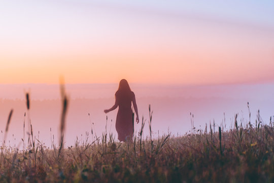 silhouette young woman in a dress walking on a hill on sunrise. female running on a foggy field and enjoying forest landscape. Active life, adventure, freedom, morning, nature concept. dew on grass.