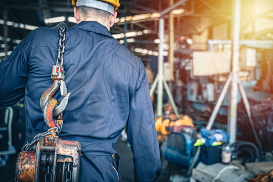 Male worker in blue jumpsuit and hardhat carrying chain hoist on his back.