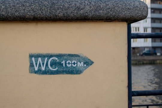 The direction sign to the toilet, inscription "WC 100 m." drawn in blue paint using a stencil on a concrete pedestal painted in beige with a marble top which is part of the fence of city embankment