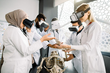 Group of young multiethnic medical students or doctors wearing VR glasses studying human skeleton anatomy in modern classroom. Medicine, anatomy, virtual reality concept
