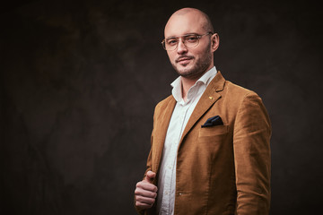 Successfull well-dressed mature bald businessman posing for camera in a dark studio wearing stylish mustard color velvet jacket, white shirt and glasses