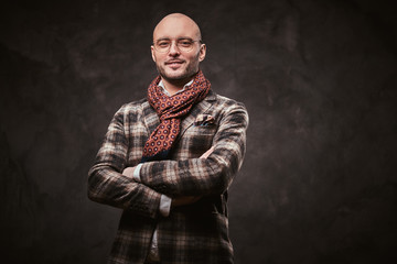 Attractive and confident stylish bald businessman posing with crossed arms for camera in a dark studio wearing checkered wool jacket, glasses and ruby patterned scarf