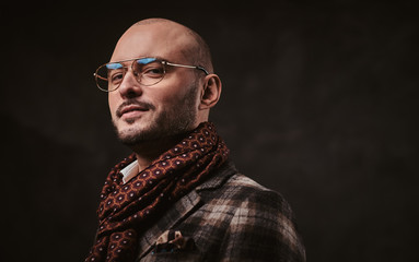 Smirking and confident stylish bald businessman posing for camera in a dark studio wearing checkered wool jacket, glasses and ruby patterned scarf