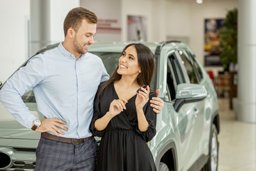 caucasian couple hugging, holding car keys at dealership salon. young couple buying their first new family automobile at car salon. lovely man and woman look at each other, woman hold car keys