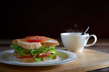 Sandwich cheese and thick ham with veg. in white dish and black coffee all put on wooden chopping board