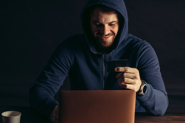 smiling caucasian hacker man in the hood of pullover sit holding bank card, going to crack...