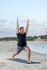 Young yoga man wearing black t-shirt at the beach, relaxing, breathing and meditating in a natural environment with palm trees and ocean as background.  - obrazy, fototapety, plakaty
