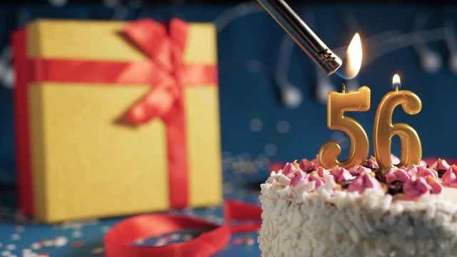 White birthday cake number 56 golden candles burning by lighter, blue background with lights and gift yellow box tied up with red ribbon. Close-up