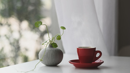 Red coffee cup with coaster putting together with ivy plant in vase decorated on modern white table with beautiful outdoors view as background.