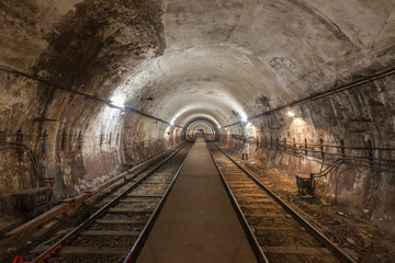 A dimly lit subway railway tunnel with an illuminated utility platform tunnel. A red light is lit...