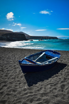 Blue boat on black sands of Ajuy beach in Fuerteventura, Canary Islands, Spain. Turquoise Atlantic ocean and black volcanic cliffs in background. Blue sky and clouds, sunny day.