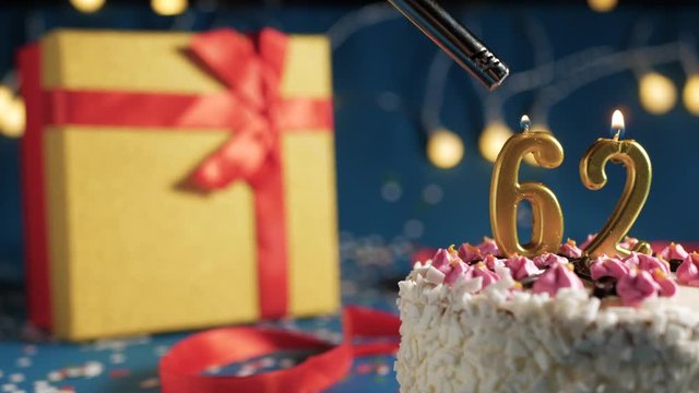 White birthday cake number 62 golden candles burning by lighter, blue background with lights and gift yellow box tied up with red ribbon. Close-up