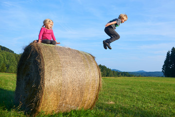 Naklejka na ściany i meble A young children boy and girl (siblings) playing on hay bale summertime. Children summer outdoor activities. Jumping high and far. Concept of summer vacation activity. Happy siblings playing on meadow
