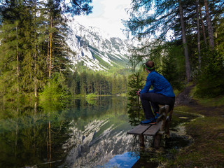Man sitting on the bench, surrounded by the water of Green Lake in an Alpine valley in Austria and enjoying the mountain view. Spring in the valley. Dense forest in the back. Playtime and happiness.