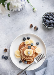 pancakes with fresh blueberries, banana on a gray background with white flowers. tasty breakfast. vertical image, top view