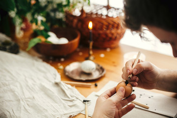 Painting Easter egg with hot wax on background of rustic wooden table with candle, basket, greenery. Easter egg with modern ornament in hand. Happy Easter. Ukrainian traditional pysanka. Zero waste