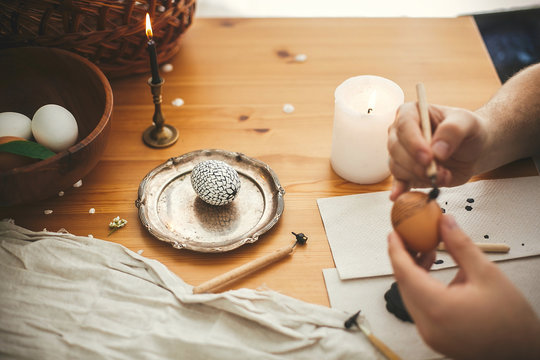 Easter egg with modern ornament  and painting Easter egg with hot wax on background of rustic wooden table with candle, basket, greenery. Happy Easter. Ukrainian traditional pysanka.