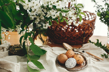 Fototapeta premium Natural Easter eggs painted with wax on vintage plate on background of wicker basket, white spring flowers and greenery on wooden table. Happy Easter. Stylish Rural still life. Zero waste