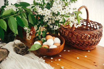 Natural Easter eggs, wicker basket, bird nest and cherry flowers on rustic table. Happy Easter, atmospheric moment. Rural still life. White and brown organic eggs. Zero waste