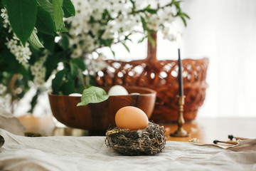 Natural Easter egg in rustic nest on background of candle, wicker basket, organic eggs, cherry flowers on wooden table. Happy Easter, atmospheric moment. Rural still life.  Zero waste