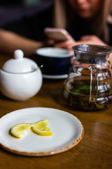 woman with a phone in the background. slices of lemon lie on white plate, next to teapot of black tea and mug and saucer