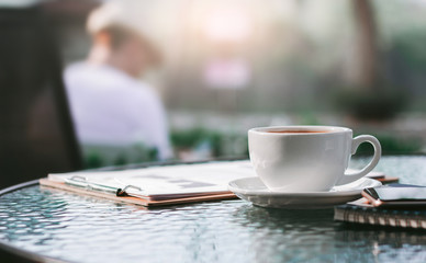 Close up white cup of coffee on a glass table with paperwork, notebook and mobile phone, Bright nature background