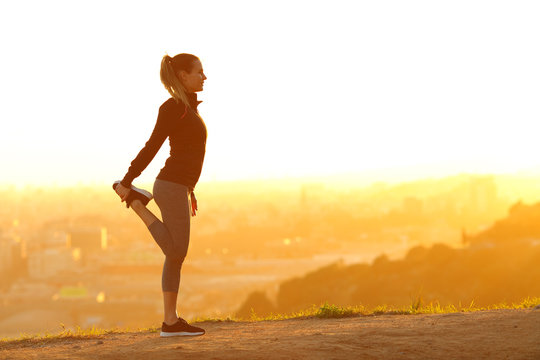 Runner woman stretching leg in city outskirts at sunset