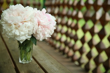 A bouquet of pink peonies stand in a glass vase against the background of a wooden table and a carved wooden gazebo