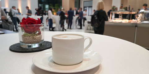 White cappuccino cup on saucer on table on blurred background with small rose arrangement in glass,many business people during coffee break at meeting,event,near tables,buffet counter on the left
