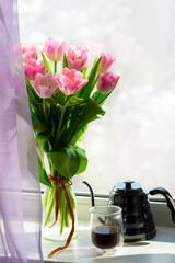 Vase with flowers on the windowsill. A cup of coffee by the window with a bouquet of flowers. Pink tulips in a vase with a teapot. March 8, Women's Day. Mother's Day. Beautiful romantic morning.