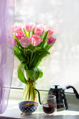 Vase with flowers on the windowsill. A cup of coffee by the window with a bouquet of flowers. Pink tulips in a vase with a teapot. March 8, Women's Day. Mother's Day. Beautiful romantic morning.