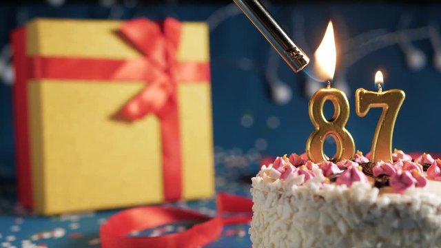 White birthday cake number 87 golden candles burning by lighter, blue background with lights and gift yellow box tied up with red ribbon. Close-up