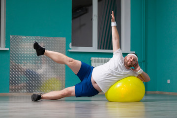 Smiling fat man is doing exercises using fitness ball. Overweight man is happy with the result of...