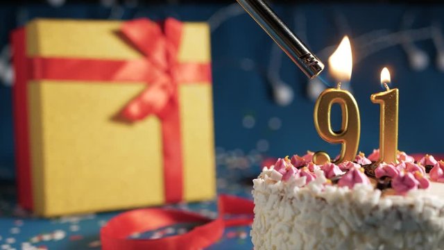 White birthday cake number 91 golden candles burning by lighter, blue background with lights and gift yellow box tied up with red ribbon. Close-up