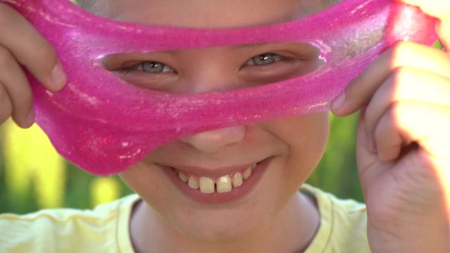 Closeup view portrait of cute funny face of white kid playing cheerfully outdoor and looking through pink circle made of transparent slime toy.