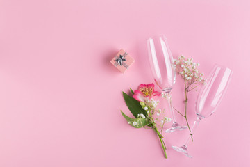 A bouquet of pink and white flowers, a small gift box and two glasses for sparkling wine on a pink paper background.