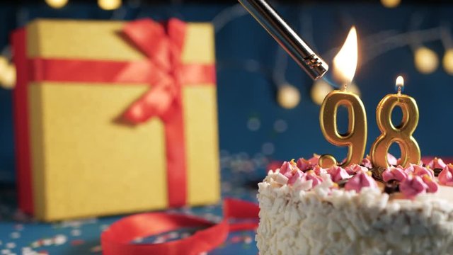 White birthday cake number 98 golden candles burning by lighter, blue background with lights and gift yellow box tied up with red ribbon. Close-up