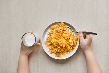Healthy breakfast with whole grain cornflakes and a glass of milk. Hands of a child holding a spoon and a glass of milk on a background of a wooden table.