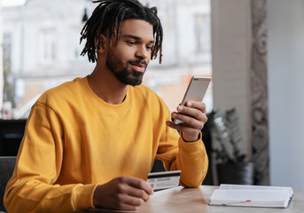 African American man holding credit card, using smartphone for online shopping, mobile banking. Young freelancer receive payment, check balance. Pensive hipster ordering food online, booking tickets