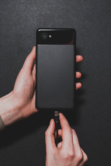 close-up of female hands that plug a black smartphone into charging on a black background