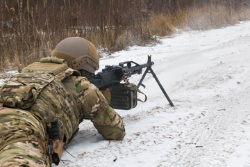 An armed soldier in camouflage uniforms lies on the snow and fires a machine gun. Winter landscape, snow field around