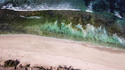 A done shot of Nyang Nyang Beach, Bali, Indonesia. The waves are rushing to the shore, making the water bubbly. The beach is covered with green algae, further on it's sandy. Idyllic hidden beach