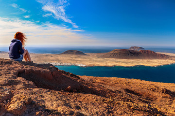 A woman looking at the sea and the island. Location: Europe, Spain, Canary Islands, Lanzarote (near Mirador del Rio; La Graciosa island in the background) - obrazy, fototapety, plakaty
