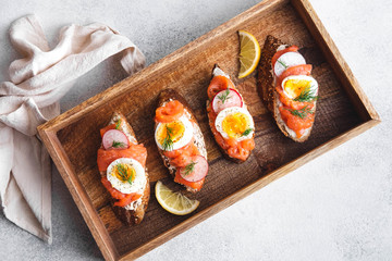 Italian appetizer bruschetta with red fish, boiled egg, cream cheese, radish and dill in a wooden box on a white background, close up top view horizontal