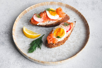 Italian appetizer bruschetta with red fish, boiled egg, cream cheese, radish and dill on a plate on a white background, close up horizontal