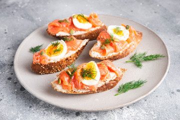 Italian appetizer bruschetta with red fish, boiled egg, cream cheese and dill on a grey plate on a grey background, close up horizontal