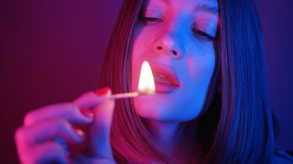 Obraz premium Attractive young woman holding matchstick with light in fingers, blowing out, looking at camera. Blue and purple combination of lights. Bob hairstyle. Red nails.