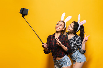 Two Young sexy caucasian woman wearing cute easter rabbit ears making selfie using stick over yellow isolated background.