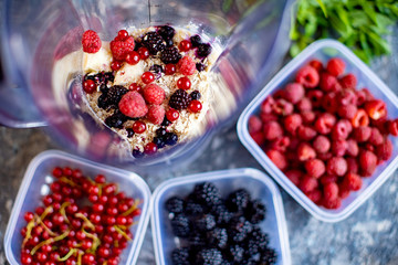 Blender with oatmeal, plastic plates, containers with raspberry, blackberry, red currant, cherry on a stone background. Making a healthy smoothie on a blender.